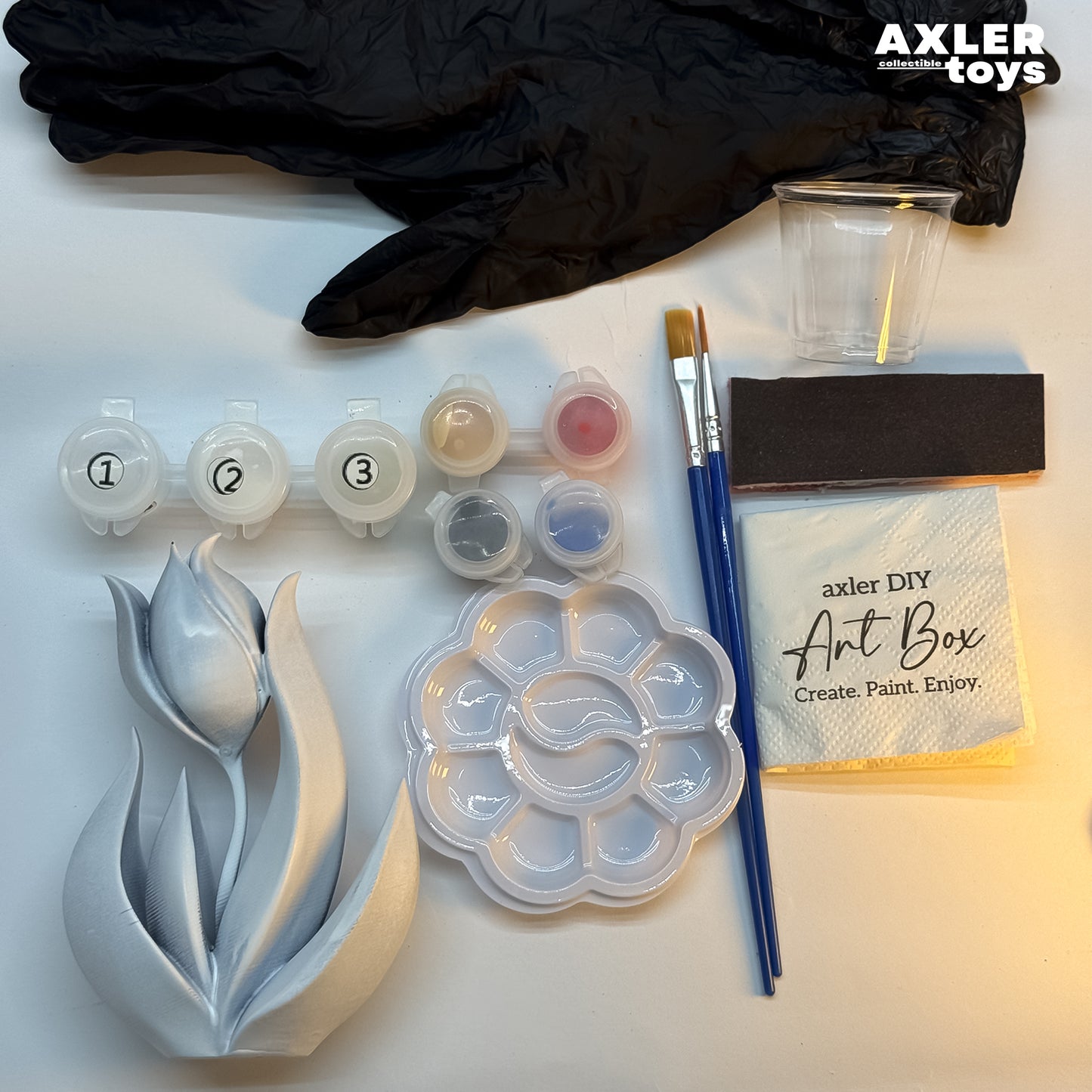 Art supplies including a tulip mold, paint containers, brushes, and a napkin with 'axler DIY Art Box' text on a white background.